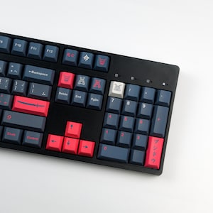 May include: A black mechanical keyboard with blue and red keycaps. The keys have Japanese characters and symbols. The number keys are red, and the function keys are blue. The keyboard is on a white surface.