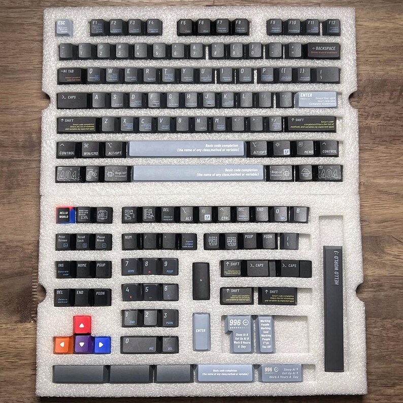 May include: A collection of black and grey keyboard keycaps, organised in a white foam insert. The keycaps have white lettering and symbols, with some keys in red, orange, and blue. Text on the keys includes "ESC", "ENTER", and "HELLO WORLD".