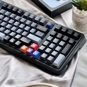 May include: A dark grey mechanical keyboard with a variety of coloured keycaps. The keyboard has a number pad and function keys. Some keycaps are blue, red, orange, and purple. The keyboard is on a grey satin surface.