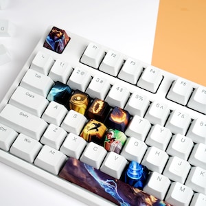 Puede incluir: Un teclado mecánico blanco con teclas personalizadas con diseños artísticos coloridos. Las teclas tienen varios patrones, incluyendo temas celestiales y de fantasía. Algunas teclas están dispersas.