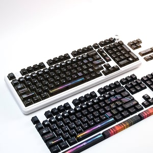 May include: Two mechanical keyboards with black keycaps and white frames are displayed. The keycaps have a gradient design. Some keycaps are detached. The keyboards are on a white surface.