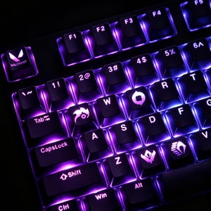 Könnte beinhalten: Eine schwarze mechanische Tastatur mit leuchtend violetter Hintergrundbeleuchtung. Die Tastenkappen zeigen Standard-Zeichen sowie benutzerdefinierte Valorant-Symbole auf den Tasten Q, E und X und das Valorant-Logo auf der Esc-Taste.
