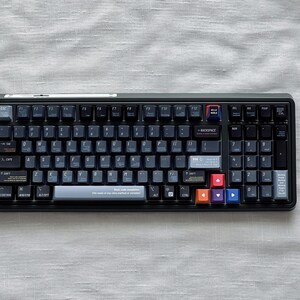May include: A black mechanical keyboard with dark grey keycaps and colourful accent keys. The keyboard is rectangular and rests on a light grey, textured surface. The keys are labelled with white lettering and symbols.