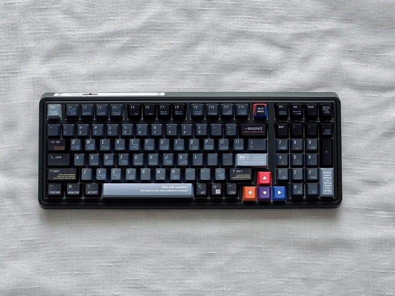 May include: A black mechanical keyboard with dark grey keycaps and colourful accent keys. The keyboard is rectangular and rests on a light grey, textured surface. The keys are labelled with white lettering and symbols.