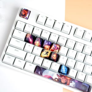 May include: White mechanical keyboard with custom keycaps featuring colorful fantasy art. The keyboard has a compact layout with a variety of illustrated keycaps. Additional white keycaps are scattered around the keyboard.
