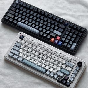 May include: Two mechanical keyboards, one black and one white, are displayed. The black keyboard has dark keycaps with colorful accent keys. The white keyboard has light-colored keycaps with blue accents. Both keyboards have a variety of keys and function keys.