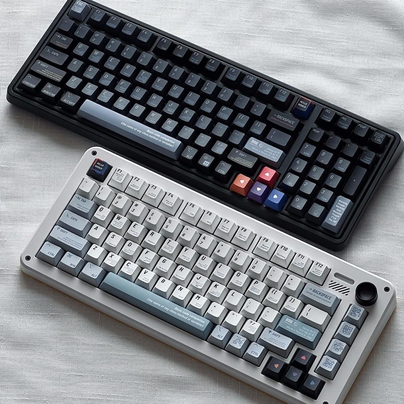 May include: Two mechanical keyboards, one black and one white, are displayed. The black keyboard has dark keycaps with colorful accent keys. The white keyboard has light-colored keycaps with blue accents. Both keyboards have a variety of keys and function keys.