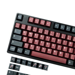 May include: A black mechanical keyboard with burgundy keycaps. The keyboard has a standard QWERTY layout with function keys and a number pad. Additional keycaps with decorative icons are included. The keys are white and burgundy.