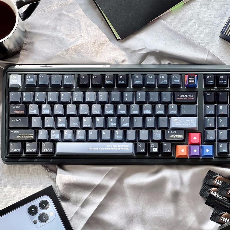 May include: A black mechanical keyboard with grey keycaps and colourful accent keys. The keyboard has a variety of function keys and a "HELLO WORLD" key. The keyboard is surrounded by a cup of coffee, a notebook, and a smartphone.