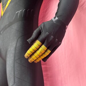 以下が含まれることがあります： 黒と黄色のスーパーヒーローコスチューム、テクスチャード加工のパターン。 手袋の指には黄色のアクセントがあります。 コスチュームは、洗練された、ぴったりとしたデザインです。