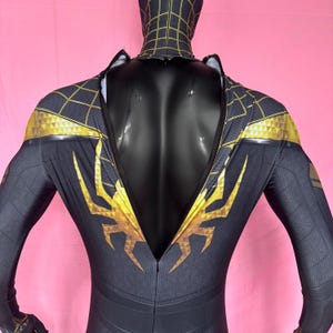 以下が含まれることがあります： 黒と金のスーパーヒーローコスチューム、フルフード付き。 スーツはウェブのようなパターンと肩と胸の金色のアクセントが特徴です。 背中にはジッパーが付いています。 コスチュームは黒いマネキンに展示されています。