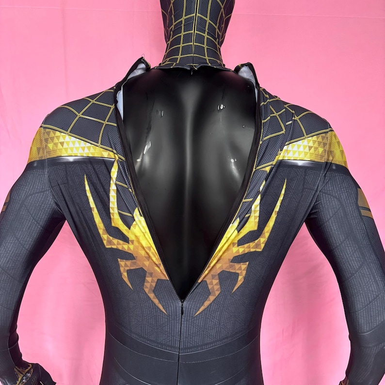 以下が含まれることがあります： 黒と金のスーパーヒーローコスチューム、フルフード付き。 スーツはウェブのようなパターンと肩と胸の金色のアクセントが特徴です。 背中にはジッパーが付いています。 コスチュームは黒いマネキンに展示されています。