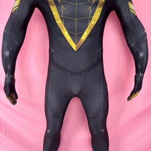 以下が含まれることがあります： 黒と金のスーパーヒーローコスチューム、テクスチャード加工。 胸と肩にV字型のゴールドデザインがあり、手袋にゴールドのアクセントが付いています。 コスチュームは体にフィットし、柔軟な素材で作られているようです。