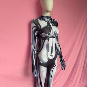 May include: A full-body jumpsuit with a high neckline and long sleeves. The jumpsuit has a black, white, and grey abstract design, creating a liquid or metallic effect. The mannequin is positioned against a pink backdrop.