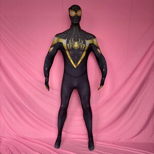以下が含まれることがあります： 胸にクモのエンブレムが付いた黒と金のスーパーヒーローのコスチューム。全身スーツは、頭と肩にクモの巣模様があり、肩と手袋に金のアクセントが付いています。コスチュームはピンク色の背景に表示されています。