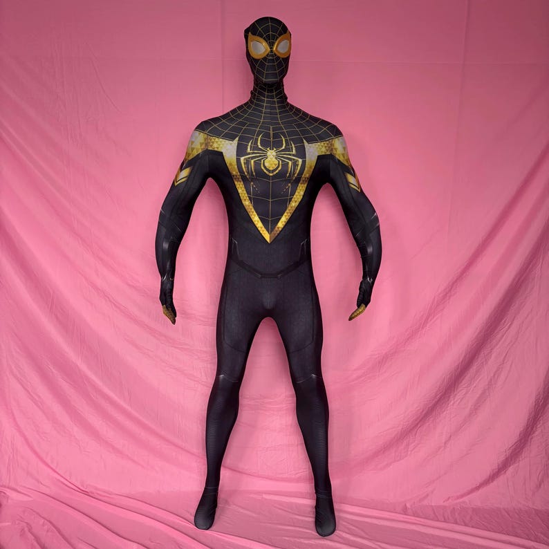 以下が含まれることがあります： 胸にクモのエンブレムが付いた黒と金のスーパーヒーローのコスチューム。全身スーツは、頭と肩にクモの巣模様があり、肩と手袋に金のアクセントが付いています。コスチュームはピンク色の背景に表示されています。