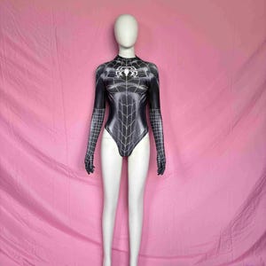 Puede incluir: Un body de manga larga en blanco y negro con un patrón de telaraña en el torso y los brazos. La prenda tiene un cuello alto y un emblema de araña blanco centrado en el pecho, con guantes integrados que cubren las manos.