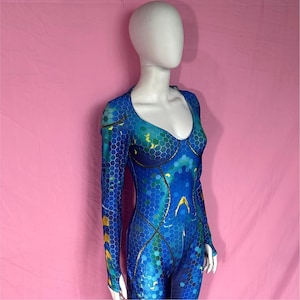 Puede incluir: Mono azul y verde con estampado de panal y mangas largas. El disfraz tiene un escote redondo y detalles dorados. La prenda se exhibe en un maniquí sobre un fondo rosa.
