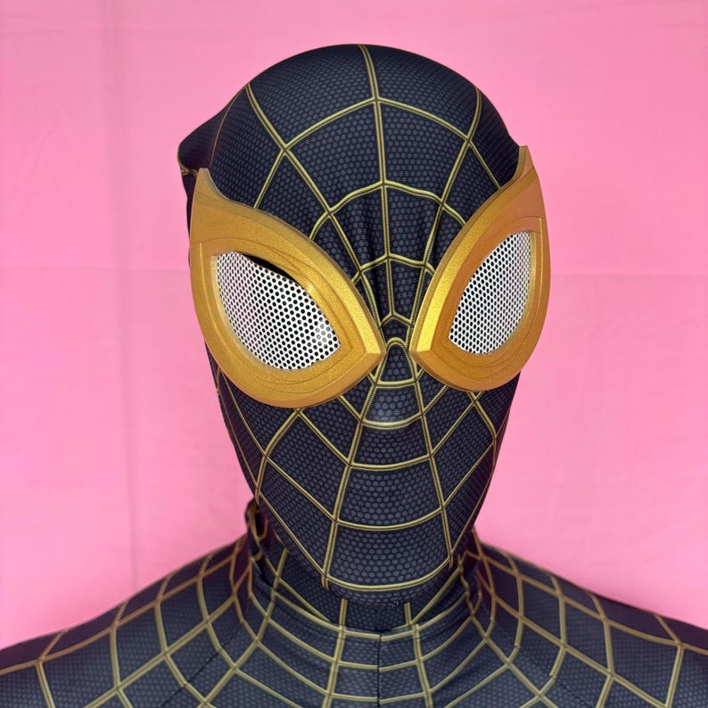 以下が含まれることがあります： 黒と金のクモの巣模様の、スパイダーマンのコスチューム。マスクは、白いメッシュの金色の縁取りのレンズが特徴です。コスチュームはピンク色の背景にあります。