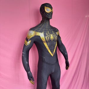 Puede incluir: Un disfraz de Spider-Man negro y dorado. El traje completo presenta un emblema de araña en el pecho y detalles dorados en los hombros, la máscara y los guantes. La máscara tiene lentes amarillas.