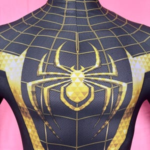 以下が含まれることがあります： 胸に金色のクモのエンブレムが付いた、黒と金のスーパーヒーローのコスチューム。コスチュームはクモの巣模様と金のアクセントが特徴です。デザインは胸と肩に見えます。