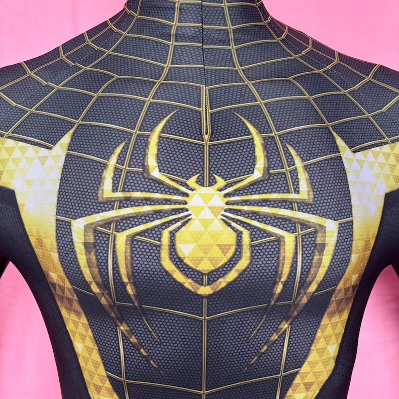 以下が含まれることがあります： 胸に金色のクモのエンブレムが付いた、黒と金のスーパーヒーローのコスチューム。コスチュームはクモの巣模様と金のアクセントが特徴です。デザインは胸と肩に見えます。