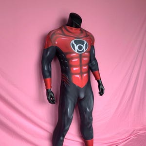 Op de afbeelding: Een rood en zwart superheldenpak met een rood borststuk en een zwart symbool op de borst. Het symbool is een cirkel met een witte omtrek en een zwarte letter "O" erin.