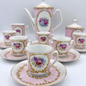 May include: A pink porcelain tea set with gold trim and floral designs. The set includes a teapot, sugar bowl, cups, and saucers. The floral design features pink, purple, and blue flowers. The set is arranged on a white surface.