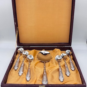 Könnte beinhalten: Ein Set aus silbernen Servierlöffeln und einer Schöpfkelle, präsentiert in einer Vintage-Box in Burgunderrot mit goldenem Innenraum. Die Löffel und die Schöpfkelle haben verzierte Griffe und eine polierte Oberfläche. Die Box ist mit einem goldenen Verschluss versehen.