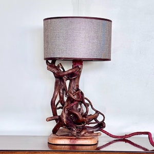 Può includere: Una lampada da tavolo con base composta da rami di legno naturale intrecciati di colore marrone scuro, montata su un basamento in legno piatto. La lampada include un paralume cilindrico in tessuto grigio testurizzato e un cavo rosso intrecciato.