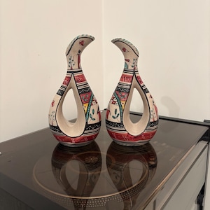 Può includere: Due vasi decorativi in ceramica con un design aperto unico. I vasi presentano una base color crema con intricati motivi dipinti a mano in rosso, nero e verde. Il design include forme geometriche e motivi stilizzati, creando un'estetica di arte popolare.