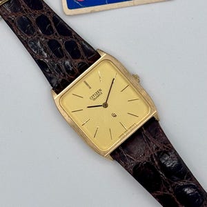Peut inclure: Une montre Citizen Quartz dorée avec un cadran rectangulaire et un bracelet en cuir marron. Le cadran affiche le logo Citizen Quartz et des repères horaires simples. Le bracelet présente un motif texturé en forme de crocodile. Une étiquette bleue et blanche est attachée.