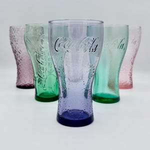 Puede incluir: Seis vasos de Coca-Cola en varios colores, rosa, verde y azul. Cada vaso tiene el logotipo de Coca-Cola y un patrón texturizado. Los vasos están colocados sobre una superficie blanca.