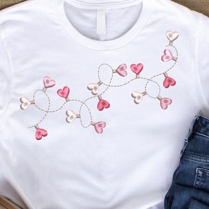 May include: White t-shirt with a pink and white heart garland design. The shirt is paired with blue jeans and brown ankle boots.