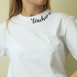 May include: White t-shirt with the text "teacher" in black cursive lettering with a heart symbol at the end. The shirt is worn by a person with their hands in their pockets. The text is located around the collar of the shirt.