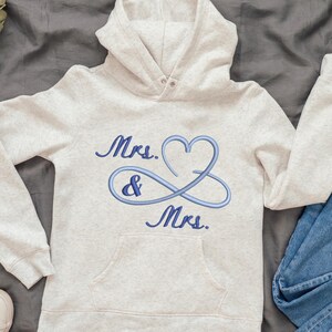 May include: A light gray hoodie with a front pocket and a hood. The hoodie features blue embroidered text and a heart-shaped infinity symbol that reads "Mrs. & Mrs.". A pair of white sneakers and blue jeans are next to the hoodie.