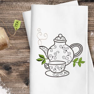 May include: White tea towel featuring a black and green embroidered design of a teapot and teacup. The teapot has a decorative swirl pattern. A tea bag and eggshells are in the background.