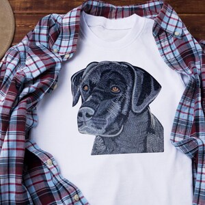 May include: White t-shirt featuring a detailed illustration of a black dog's face with brown eyes. The shirt is paired with a red, blue, and white plaid button-down shirt and a brown cowboy hat. The dog illustration is the focal point.