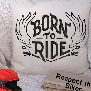 Può includere: Cuscino quadrato color crema con la scritta "BORN TO RIDE" in nero, affiancata da ali stilizzate. Un casco da moto rosso e una pila di libri con la scritta "Respect the Biker" sono anch'essi visibili.