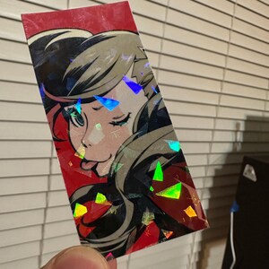 May include: A holographic sticker featuring a cartoon character with black hair, red eyes, and a red and black outfit. The character is winking and sticking out their tongue. The sticker has a red background with a holographic effect.
