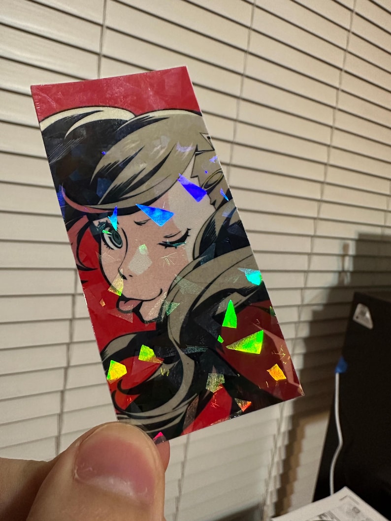 May include: A holographic sticker featuring a cartoon character with black hair, red eyes, and a red and black outfit. The character is winking and sticking out their tongue. The sticker has a red background with a holographic effect.