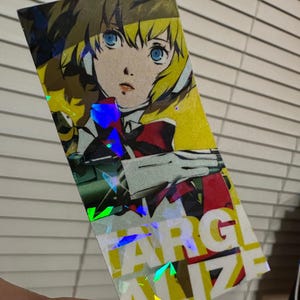 May include: A holographic sticker featuring an anime character with blonde hair and blue eyes. The character is wearing a red and white outfit and is holding a weapon. The sticker has a holographic effect with rainbow colors. The text "FARGALIZE" is visible on the sticker.