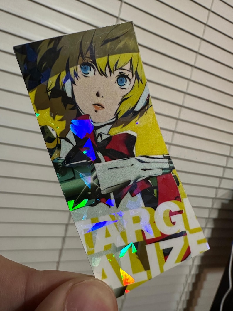 May include: A holographic sticker featuring an anime character with blonde hair and blue eyes. The character is wearing a red and white outfit and is holding a weapon. The sticker has a holographic effect with rainbow colors. The text "FARGALIZE" is visible on the sticker.