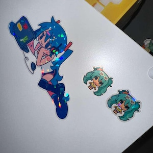 May include: Holographic stickers featuring anime-style characters. One sticker depicts a blue-haired character holding a device, while two smaller stickers show a pixelated character with green hair and sunglasses. The stickers are on a white surface.