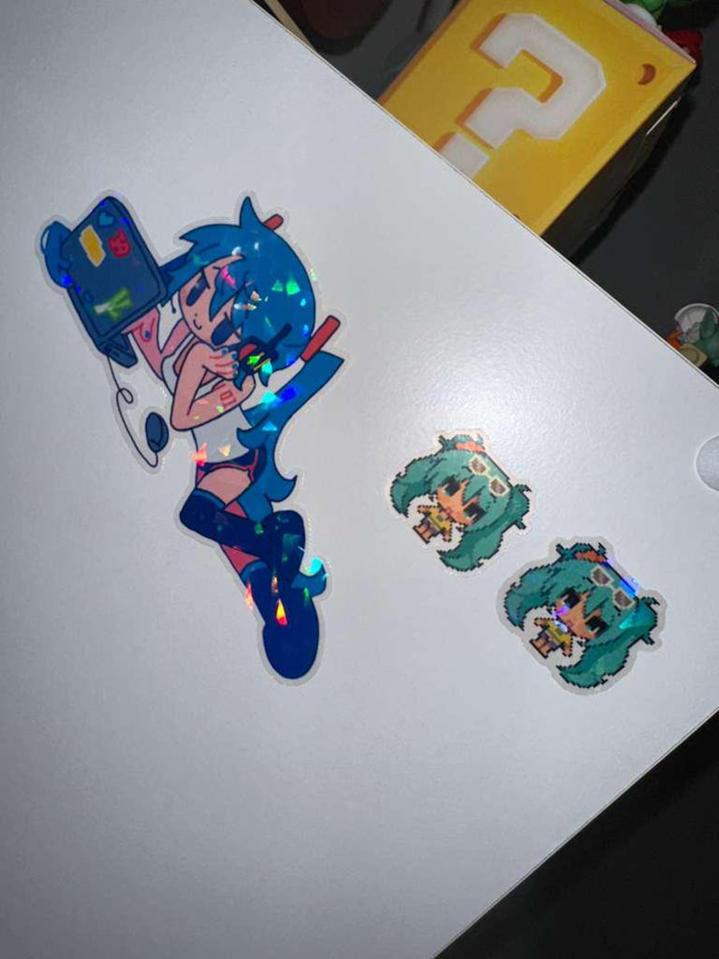May include: Holographic stickers featuring anime-style characters. One sticker depicts a blue-haired character holding a device, while two smaller stickers show a pixelated character with green hair and sunglasses. The stickers are on a white surface.