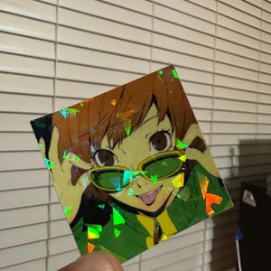 May include: A holographic sticker featuring an anime character with brown hair, wearing green sunglasses and a green and yellow shirt. The character is smiling and sticking out their tongue.