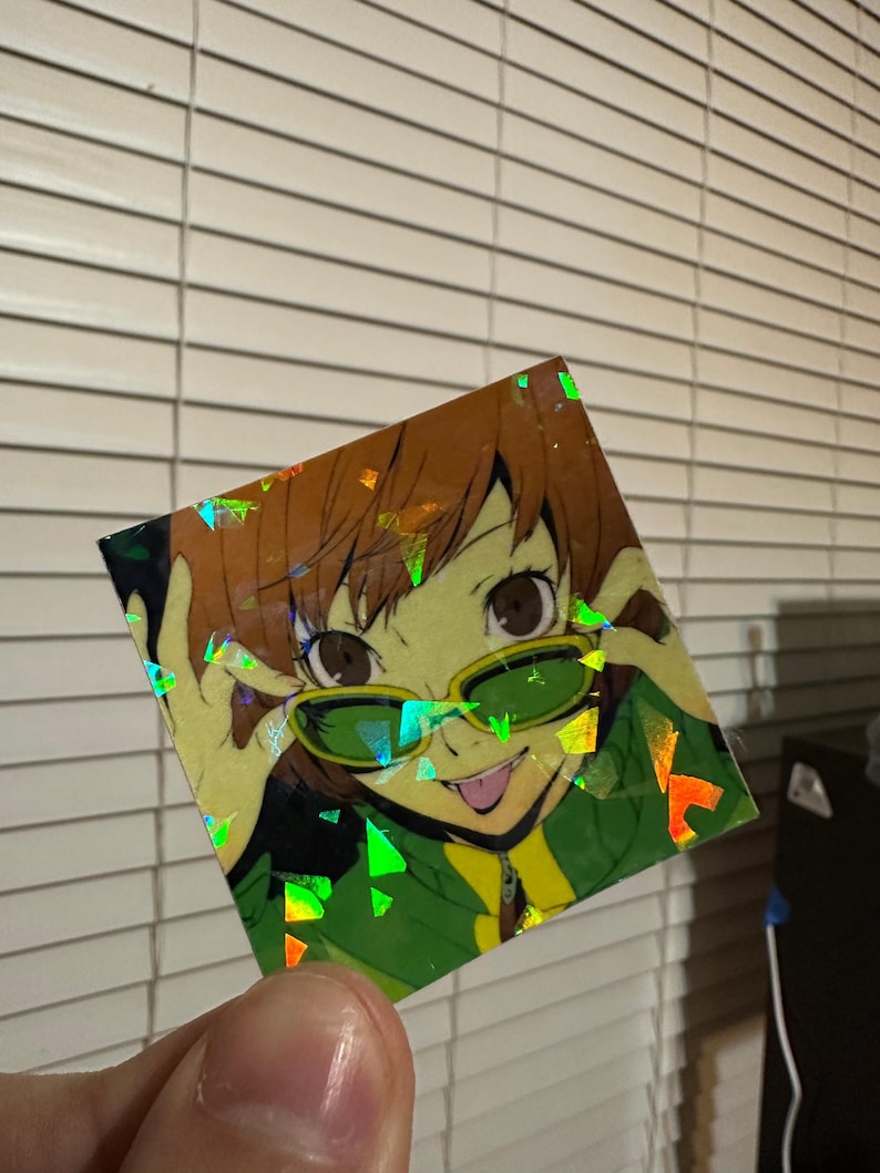May include: A holographic sticker featuring an anime character with brown hair, wearing green sunglasses and a green and yellow shirt. The character is smiling and sticking out their tongue.
