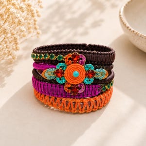 May include: A multi-layered bracelet featuring woven bands in dark brown, magenta, and orange. The centerpiece is a floral-shaped ornament with a central orange coil, a small turquoise bead, and surrounding red and blue crystals, plus green accents.
