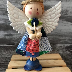 May include: A wooden angel figurine with a painted face, orange yarn hair, and a halo. It features white glittery wings, a blue fabric flower, and a patchwork dress made of blue, red, and white floral and checkered fabric, standing on a blue base.