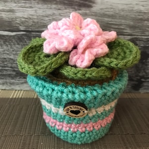 May include: A crocheted flower pot with a pink flower and green leaves. The pot is turquoise, white, and pink with a wooden button that says "Love N Stitches". The pot is sitting on a brown surface.